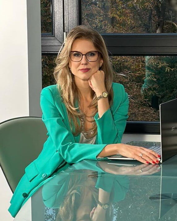 “Lawyer Klimova Anastasia in a teal suit, white blouse, and glasses, sits at a glass desk in an office, with a camera on a tripod in the foreground. A laptop and books are on the desk.”